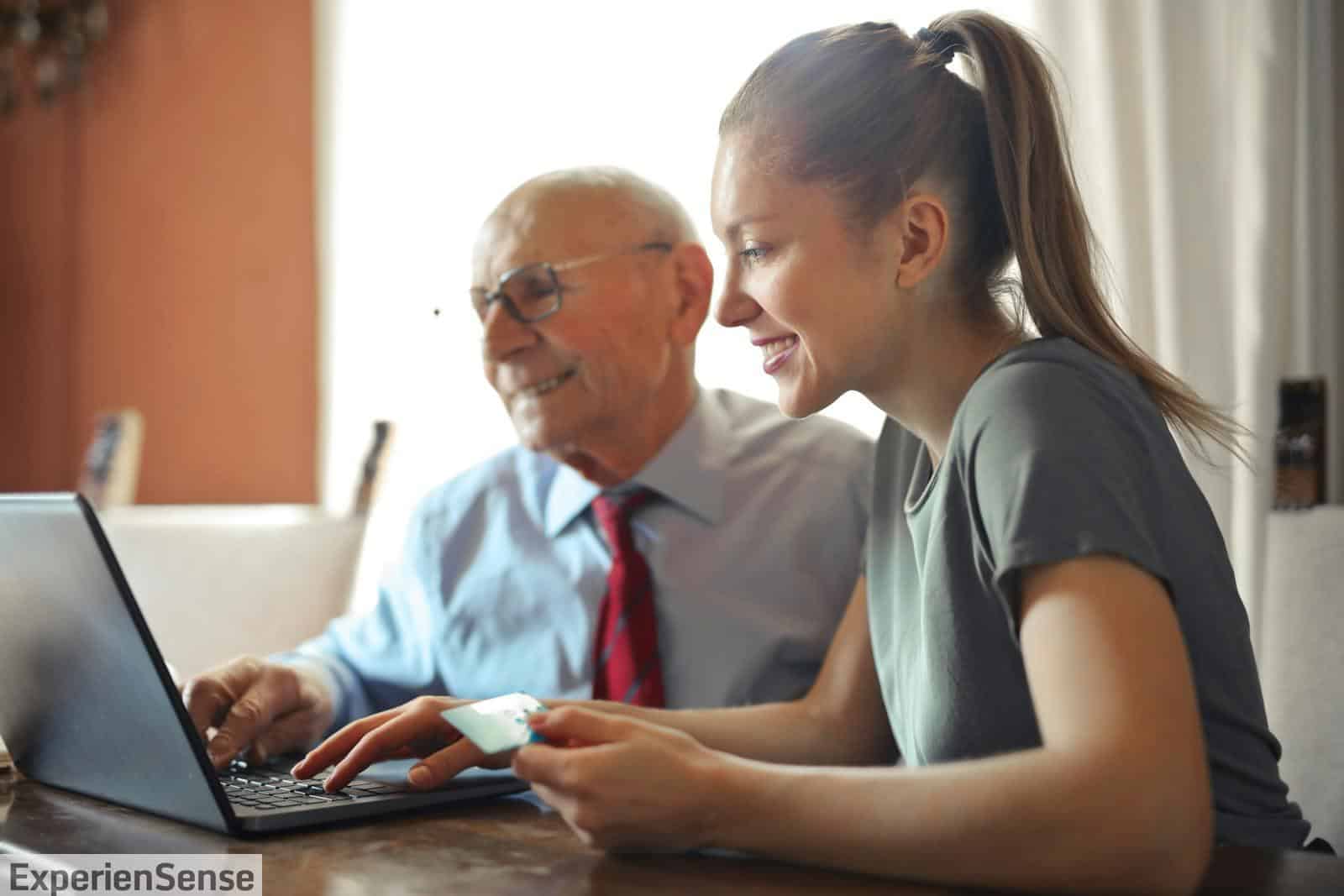 Young woman in casual clothes helping senior man in formal shirt with paying credit card in Internet using laptop while sitting at table